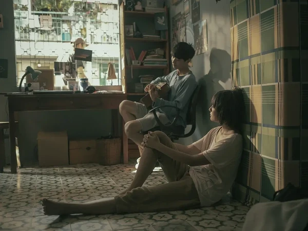 In the movie SaiGon trong cơn mưa, a guy plays guitar while a girl sits on the floor, enjoying a cozy moment.