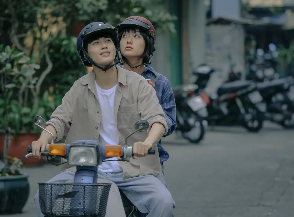 In SaiGon trong cơn mưa, a young couple takes a sweet motorbike ride, wearing helmets and looking ahead.